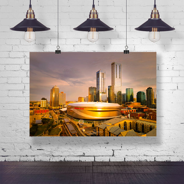 A rectangular wall art print showing a city skyline with a large stadium at sunset, hanging from three black pendant lights against a white brick wall.