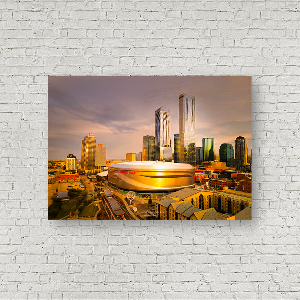 A rectangular wall art print showing a Edmonton city skyline at sunset, mounted on a white painted brick wall. The print features Rogers Place arena in the foreground with tall modern skyscrapers and warm-toned buildings in the background under a pinkish-orange sky.