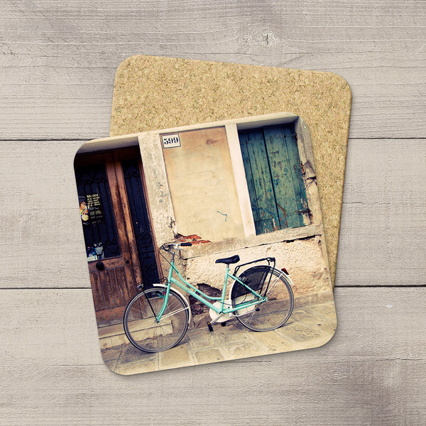 Two rectangular cork coasters with rounded corners, one featuring a printed photograph of a mint green bicycle parked in front of a rustic building with a blue door, displayed on a light gray wood plank surface.