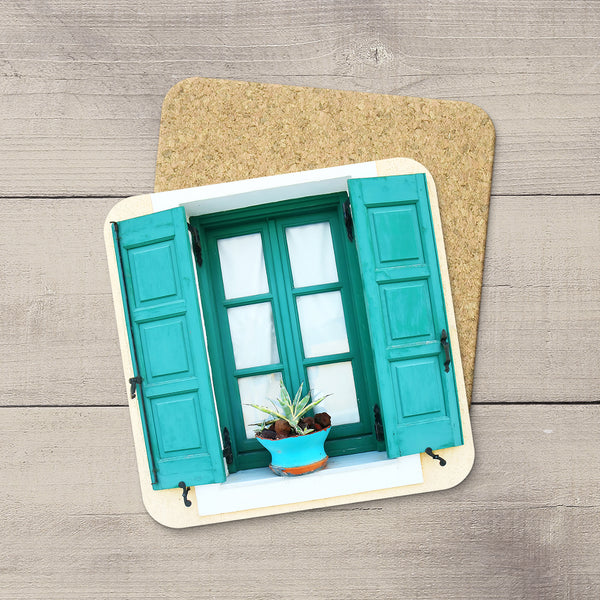 A square cork coaster featuring a printed design of a window with teal shutters and a small potted plant in Santorini, Greece, displayed on a gray wood plank surface.