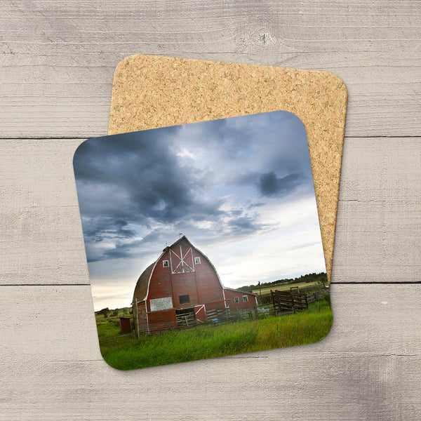 Square coaster with a red barn photo, glossy finish, and non-slip cork backing on a rustic wooden surface.