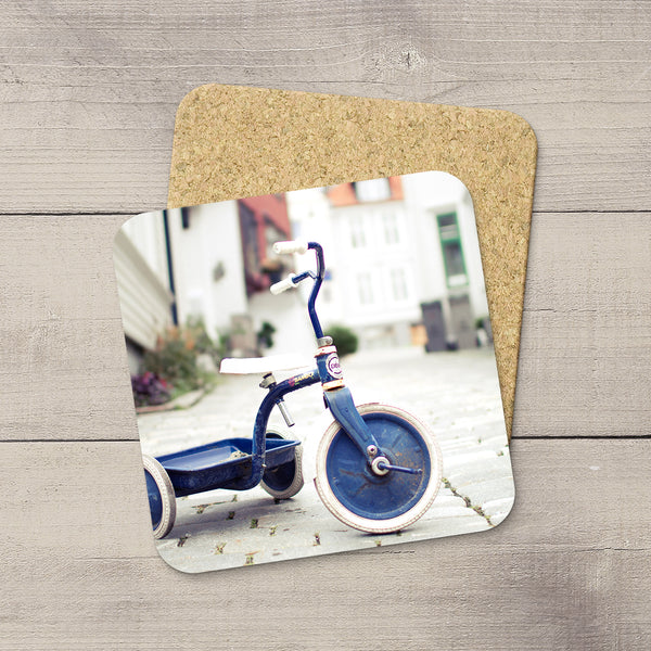 A square cork coaster with rounded corners featuring a printed photo of a blue tricycle, displayed on a light gray wood plank surface.