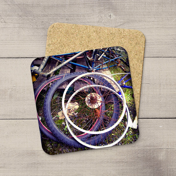 Two square coasters with rounded corners, one featuring a printed graphic of bicycle wheels and the other showing a natural cork texture, displayed on a gray wood plank background.