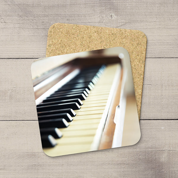A square cork coaster with rounded corners printed with a close-up photograph of piano keys, shown on a light gray wood plank surface with a second coaster underneath.