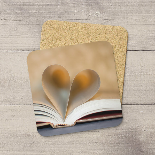 A square coaster with rounded corners featuring a printed design of an open book with heart-shaped pages, placed on top of a cork backing over a light gray wood plank surface.