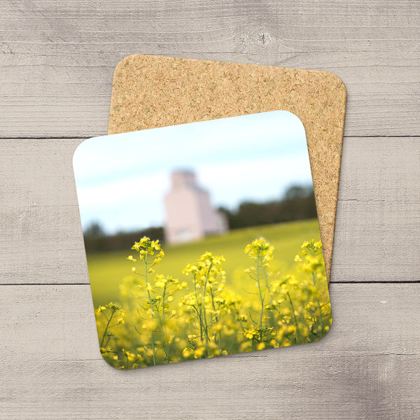 Square coaster with a glossy canola field photo, rounded corners, and a non-slip cork base on a rustic wood background.