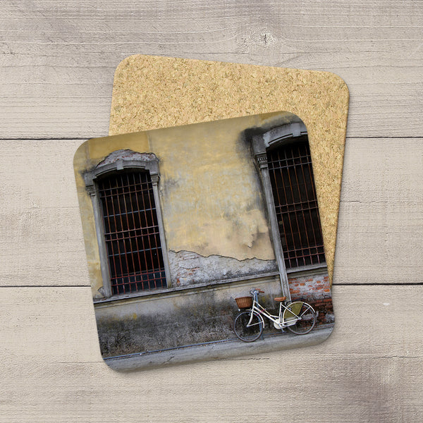 Two square cork coasters stacked on a light wood surface. The top coaster features a printed photograph of a weathered yellow building facade in Lucca, Italy with barred windows and a white bicycle leaning against the wall.