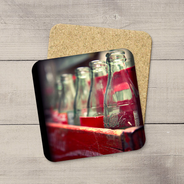A square cork coaster with rounded corners featuring a printed photograph of vintage glass Coca-Cola bottles on a red surface, displayed on a light gray wood plank background.