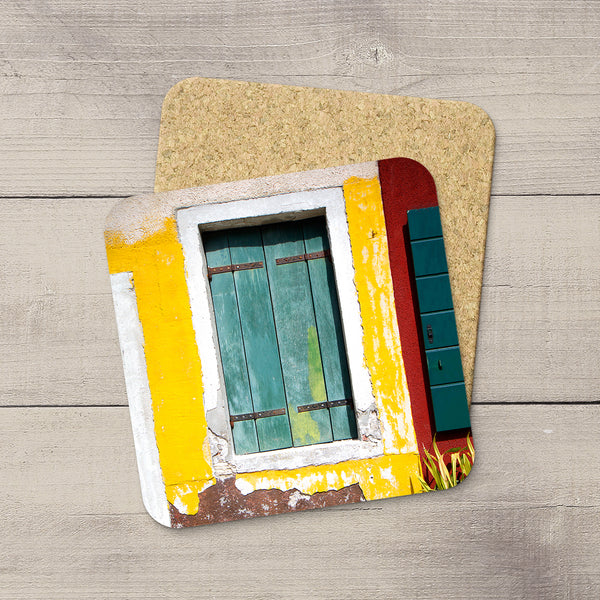 A stack of square cork coasters featuring a colorful printed design of a rustic window with yellow, teal, red, and white details, displayed on a light gray wood plank surface.