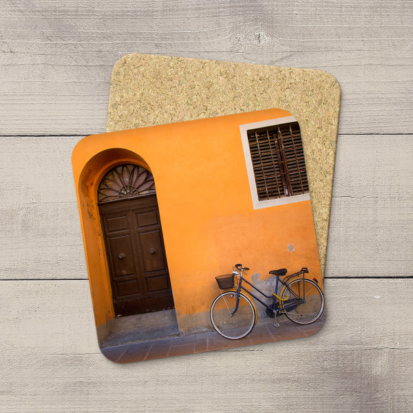 Two square cork coasters with rounded corners, one featuring a printed photograph of an orange wall with a dark wooden door and a bicycle in Pisa, Italy, displayed on a light gray wood plank background.