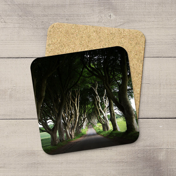 Two square cork coasters with rounded corners, one featuring a printed photograph of a tree-lined path called the Dark Hedges in Ireland, and the other showing the natural cork backing, displayed on a light gray wood surface.