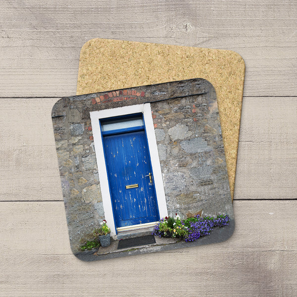 A square coaster featuring a printed photograph of a blue door on a stone wall, shown on a light wood surface with a second coaster partially visible behind it.