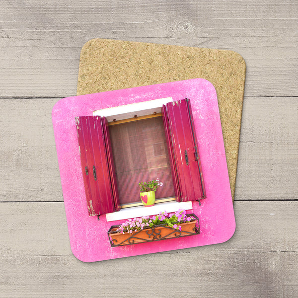 A square pink coaster with rounded corners featuring a printed window design with red shutters and a flower box, placed on top of a cork coaster over a gray wood plank surface.