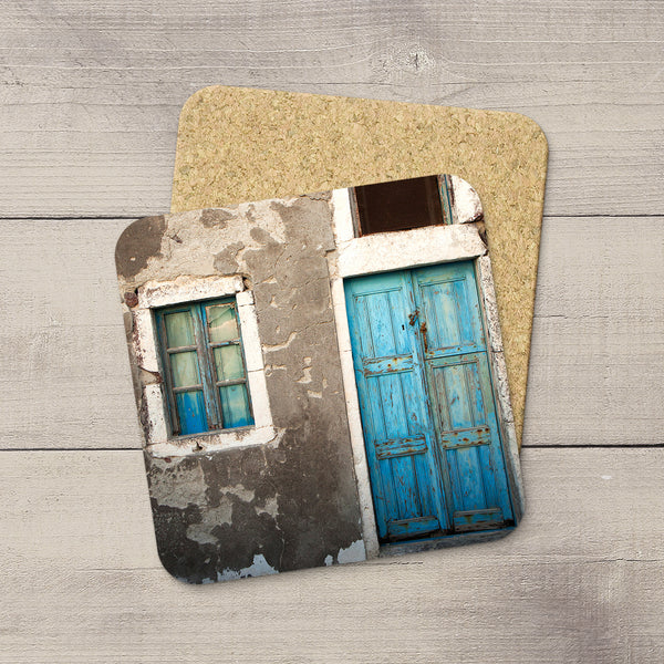 A square coaster with rounded corners featuring a printed photograph of a weathered gray wall with a bright blue door and windowin Oia, Greece, placed on top of a cork backing over a light gray wood plank surface.