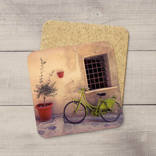 Two square coasters with rounded corners displayed on a light gray wood background. The top coaster features a printed scene of a green bicycle parked against a beige wall in Cinque Terre Italy next to a potted plant, while the bottom coaster shows the natural cork backing.