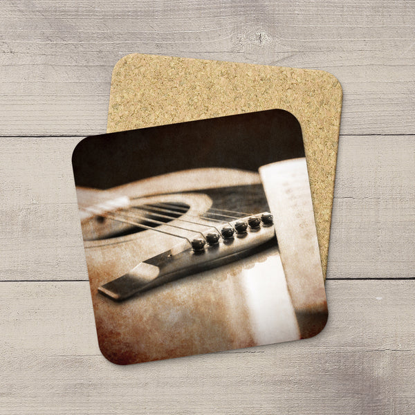 Two square coasters with rounded corners stacked on a light gray wood plank surface. The front coaster features a sepia-toned printed photograph of an acoustic guitar close-up, while the back coaster shows a natural cork texture.