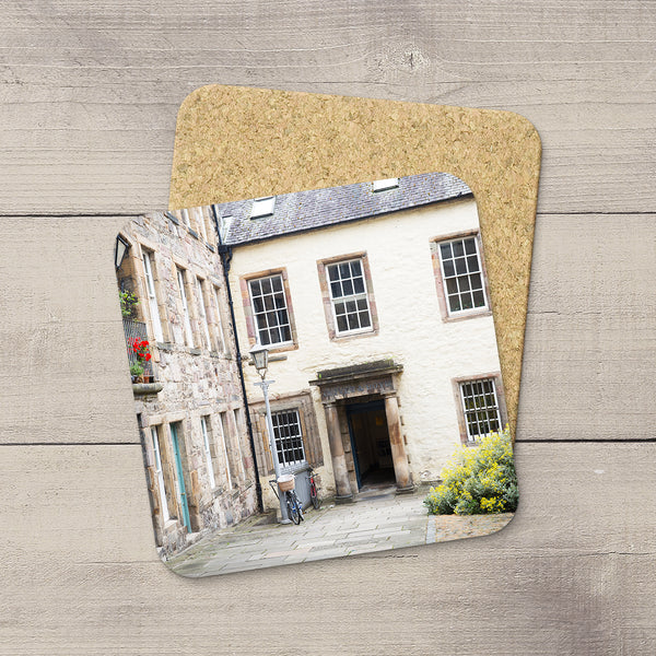 A square cork coaster with rounded corners printed with a photograph of a stone building courtyard in Edinburgh, Scotland, displayed on a light gray wood plank surface.
