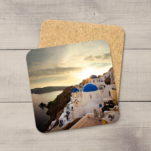 Two square cork coasters with rounded corners, one printed with a scenic photograph of Santorini featuring white buildings with blue domes and a sunset over the sea, displayed on a light gray wood background.