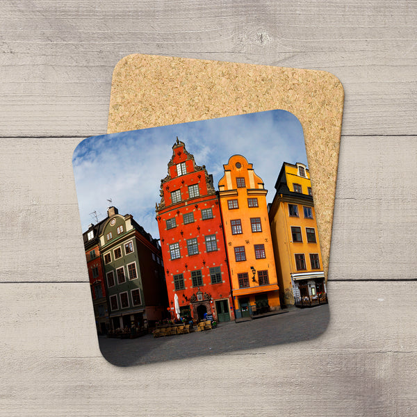 A square cork coaster with rounded corners featuring a printed photograph of colorful historic buildings in a town square of Stockholm, Sweden, displayed on a light gray wood plank surface.