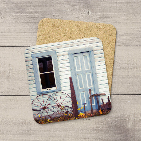 A square cork coaster with rounded corners featuring a printed photograph of a light blue rustic house exterior in Yukon, Canada, displayed on a gray wood plank surface.