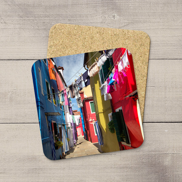 Two square coasters with rounded corners, one featuring a colorful photograph of a European alleyway with laundry lines and the other showing a cork backing, displayed on a light gray wood background.