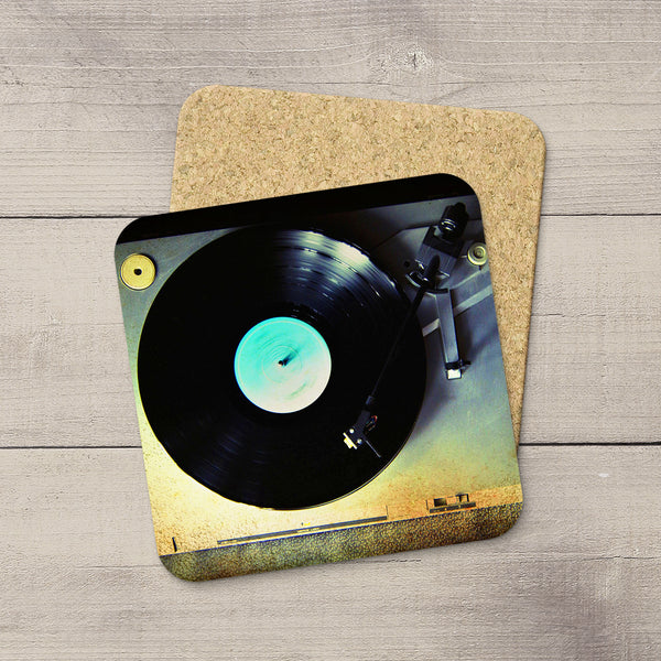A square cork coaster with rounded corners featuring a printed vinyl record and turntable design, placed on a light gray wood plank surface.