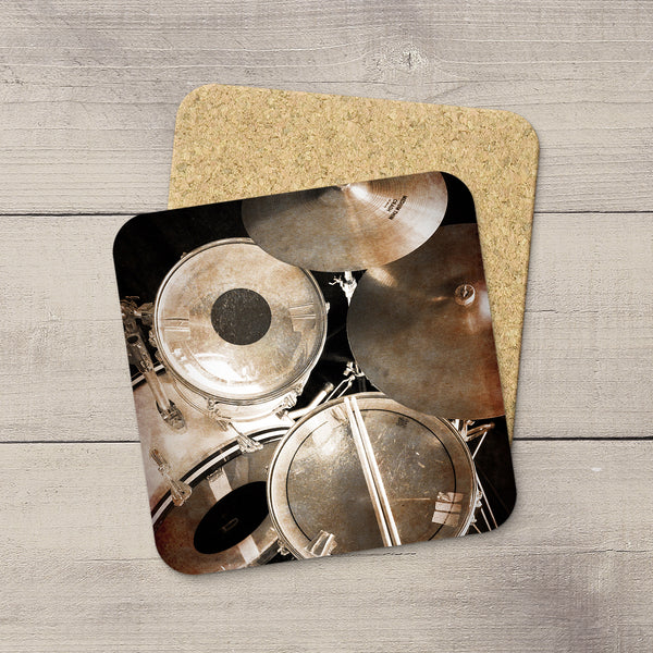 A square coaster with rounded corners featuring a sepia-toned drum set photograph, placed on top of a cork coaster over a light gray wood plank background.