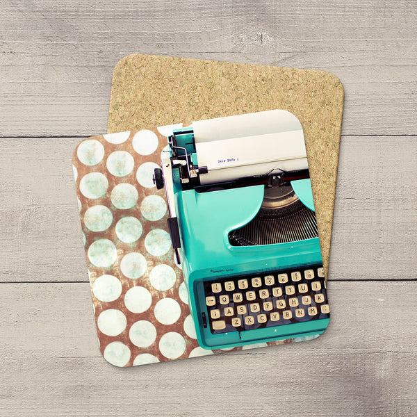 A square desk mat featuring a retro turquoise typewriter design on one side and a brown background with white polka dots on the other, displayed on a cork board over a gray wood plank surface.