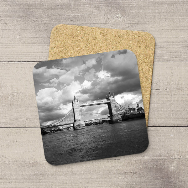 A square cork coaster with rounded corners featuring a black-and-white photograph of Tower Bridge over the River Thames, displayed on a light wood surface with a second coaster showing the natural cork backing.