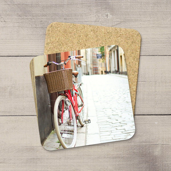 Two square cork coasters with rounded corners, one featuring a printed photo of a red bicycle on a cobblestone street and the other plain, displayed on a gray wood plank background.