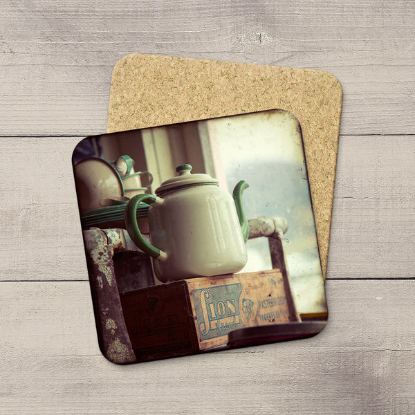 Two square cork coasters with rounded corners, one featuring a vintage-style printed photo of a cream enamel kettle on a rustic table, displayed on a light gray wood plank background.