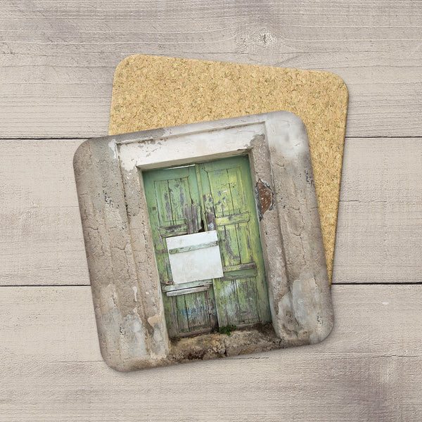 A square cork coaster featuring a printed design of a weathered green door in a rustic stone frame, displayed on a light gray wood plank background with a second coaster partially visible behind it.