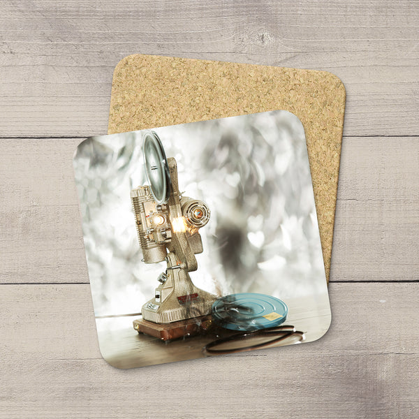 A square printed coaster with rounded corners lies on a cork backing over a light wood surface. The printed design shows a vintage film projector with a glowing bulb and a blue reel.