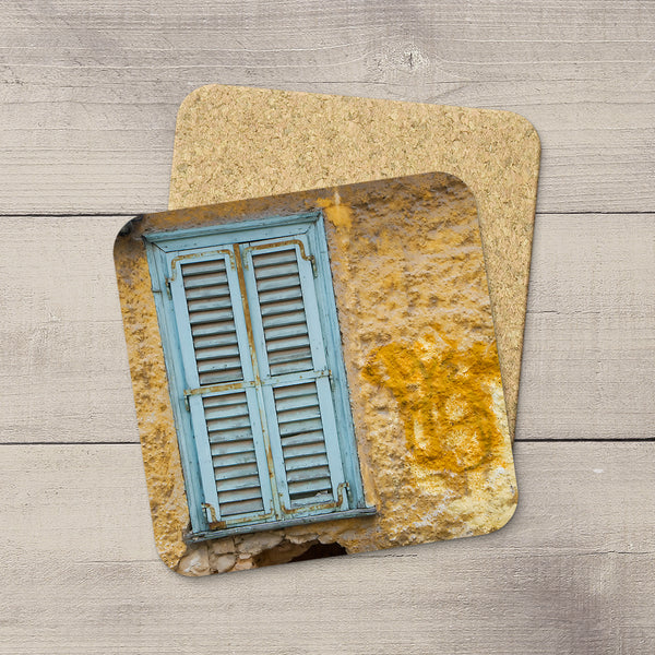 Two square cork coasters stacked on a light gray wood plank surface. The top coaster features a printed design of a rustic stone wall with a weathered light blue shuttered window and a patch of orange moss.