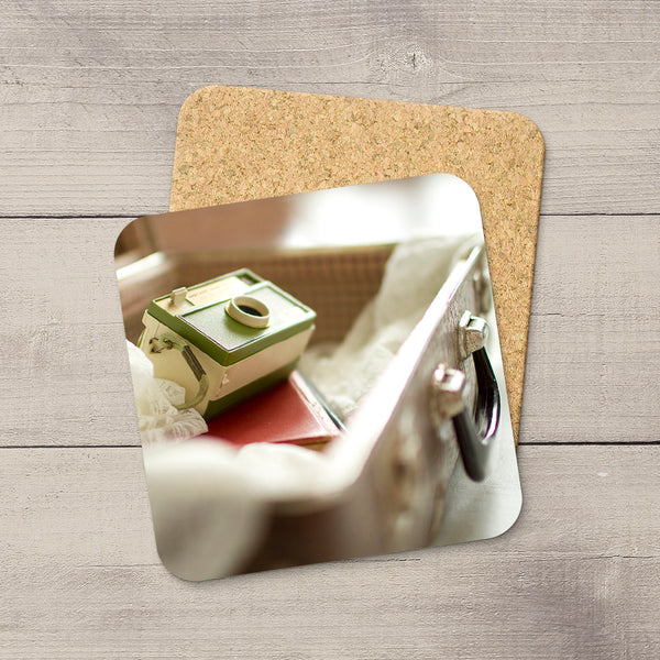 A square cork coaster with rounded corners lies on a light gray wood plank surface, partially covering a printed photo coaster. The photo coaster shows a close-up scene with a green and cream box, a red item, and a metallic handle.