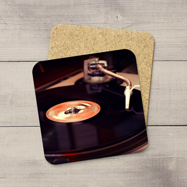 Two square coasters with rounded corners, one featuring a printed photo of a turntable and vinyl record, and the other showing a cork-like textured surface, displayed on a gray wood plank background.