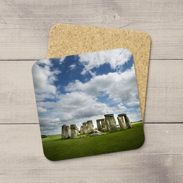 A square cork coaster with rounded corners printed with a scenic photograph of Stonehenge under a blue sky, displayed on a light gray wood surface with another coaster partially visible behind it.