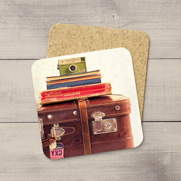 A square coaster with rounded corners featuring a vintage travel illustration of stacked books and a brown leather suitcase, placed on top of a cork square over a gray wood plank background.