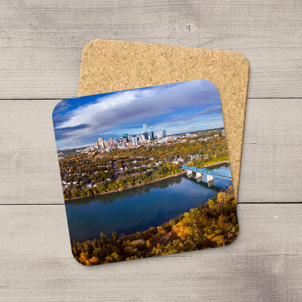 Two square cork coasters with rounded corners, one featuring a printed aerial photograph of Edmonton city skyline, river, and autumn trees, displayed on a light gray wood plank surface.
