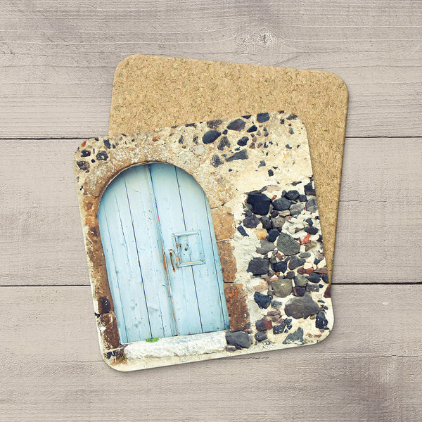 A square cork coaster featuring a printed design of a light blue wooden door set in a rustic stone wall, placed on top of a second plain cork coaster on a gray wood plank surface.