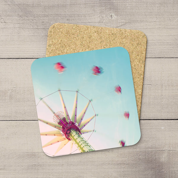 A square printed coaster with rounded corners lies on top of a cork coaster on a light gray wood plank surface. The printed side shows a whimsical photograph of an amusement ride against a pale blue sky, with pink and red ride cars blurred in motion.