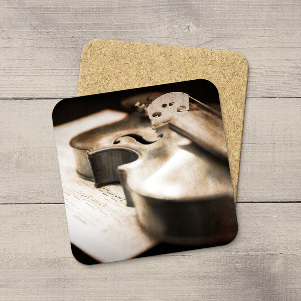 A square coaster with rounded corners featuring a sepia-toned close-up photo of a violin over sheet music, placed on top of a cork-backed coaster on a light gray wood surface.