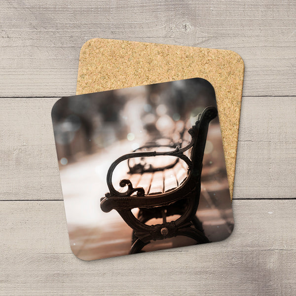 Two square coasters with rounded corners stacked on a light gray wood surface. The front coaster features a printed photograph of an empty park bench in Victoria Promenade in Edmonton, while the back coaster shows a natural cork backing.