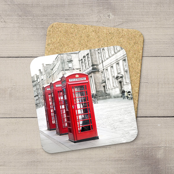 A square cork coaster with rounded corners featuring a printed photograph of classic red London telephone booths on a street, displayed on a light gray wood plank surface.