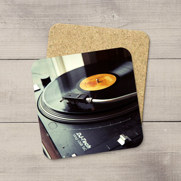 A square cork coaster with rounded corners featuring a printed photo of a turntable playing a vinyl record, placed on a light gray wood plank surface.