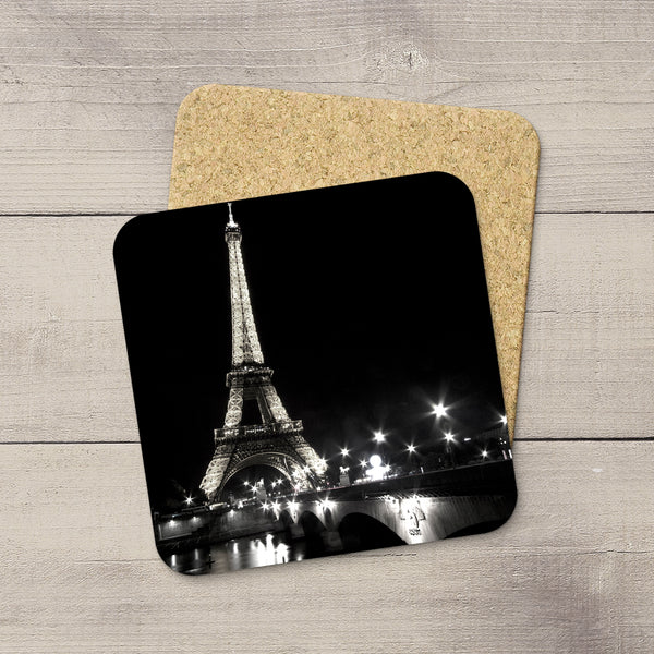 A square cork coaster with rounded corners printed with a black-and-white nighttime photograph of the Eiffel Tower, placed on a light wood surface with a second coaster partially visible behind it.