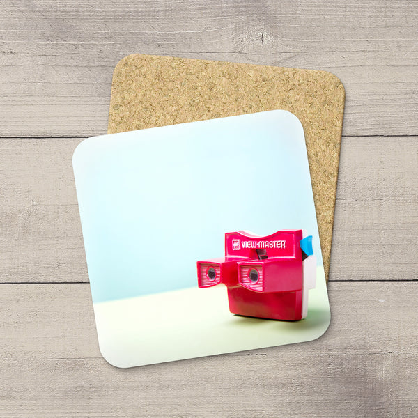 A square printed coaster or mat with rounded corners lies on a light wood surface, partially covering a cork backing. The printed design shows a bright pink ViewMaster camera against a pale blue and white background.