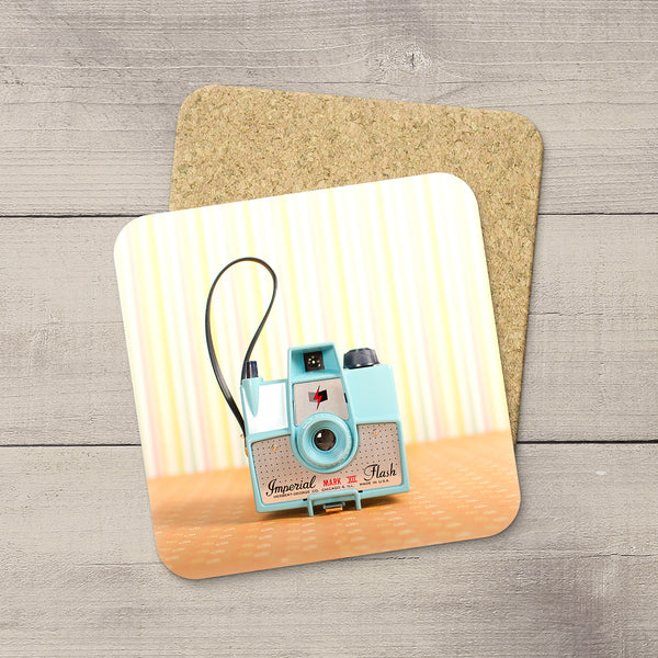 A square coaster with rounded corners featuring a vintage light blue camera graphic on a cream and yellow striped background, placed on top of a cork coaster over a gray wood plank surface.