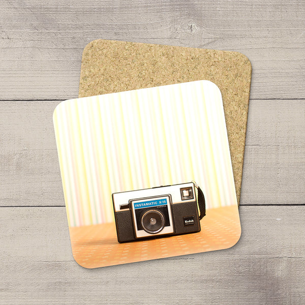 A square coaster with rounded corners featuring a retro film camera design, displayed on a cork backing over a gray wood plank surface.