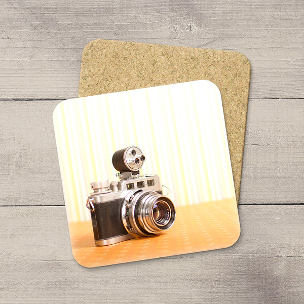 A square coaster with rounded corners featuring a printed vintage camera design, displayed on top of a second coaster showing the cork backing, set against a gray wood plank background.